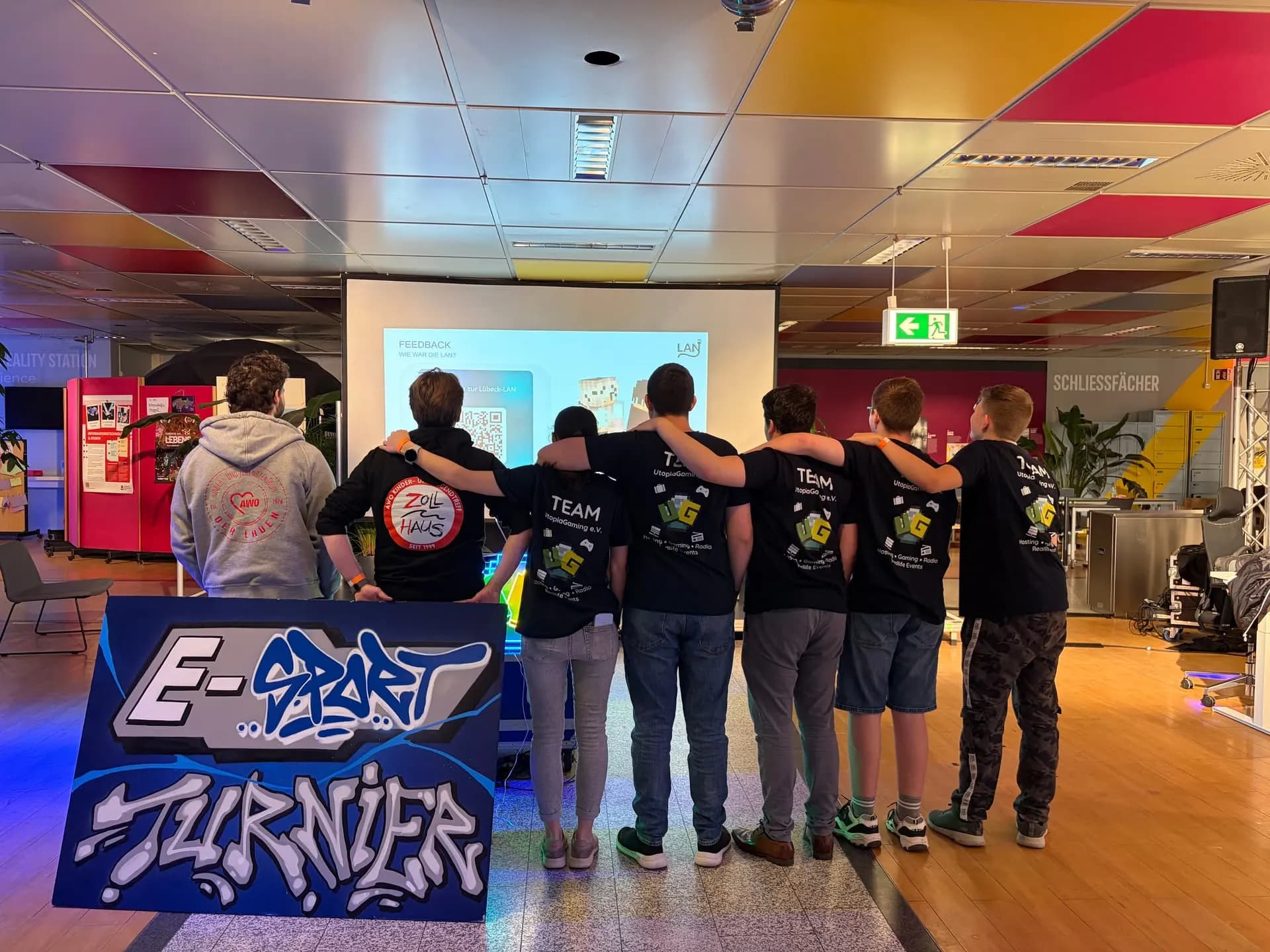Das ehrenamtliche Team der Lübeck LAN Arm in Arm vor der Feedback-Leinwand, davor ein gemaltes E-Sport-Turnier-Schild